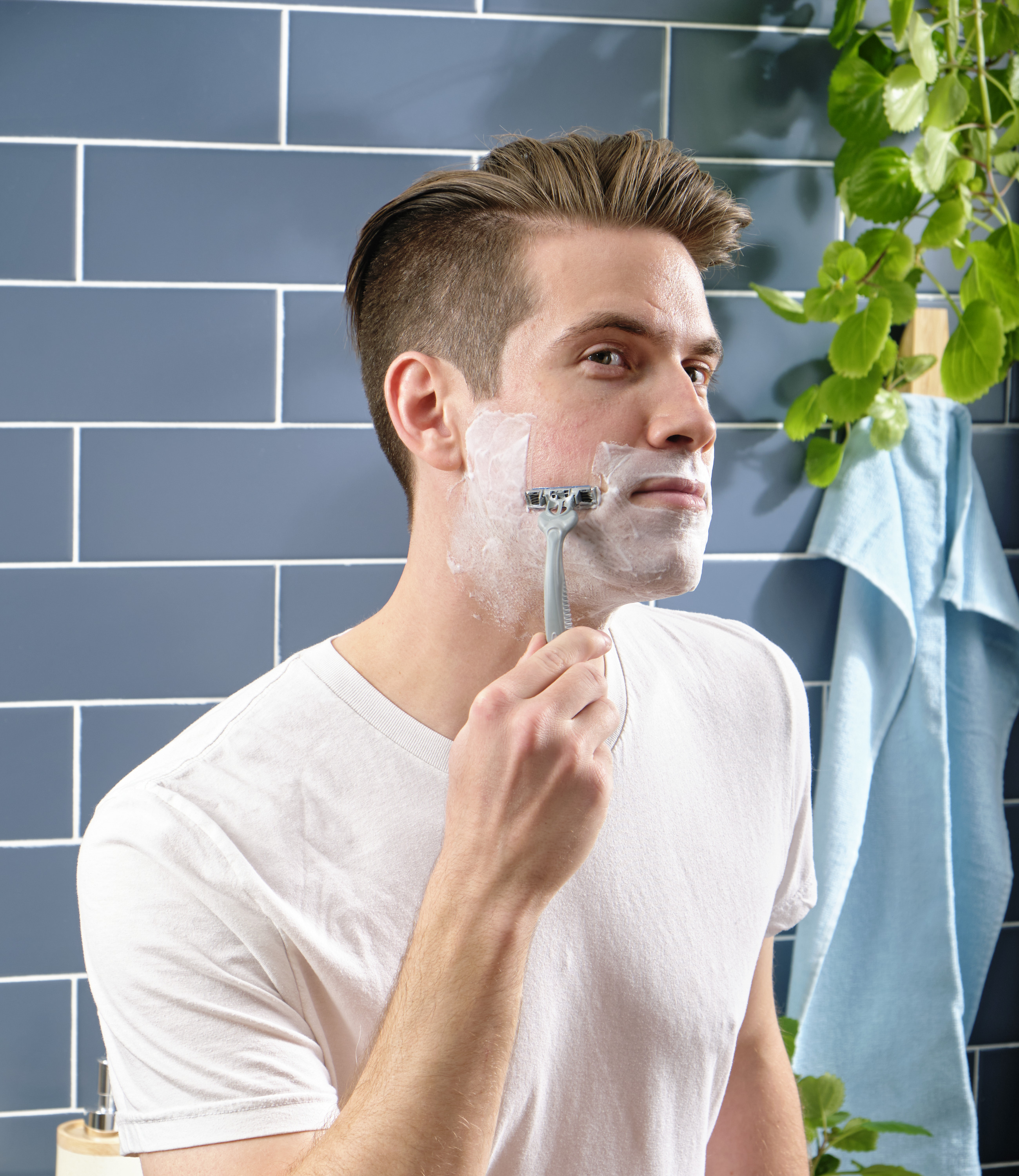 man shaving with a blue razor in front of a blue tiled wall